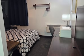 Quarto de hotel pequeno com uma cama, escrivaninha, cadeira, abajur e radiador.
