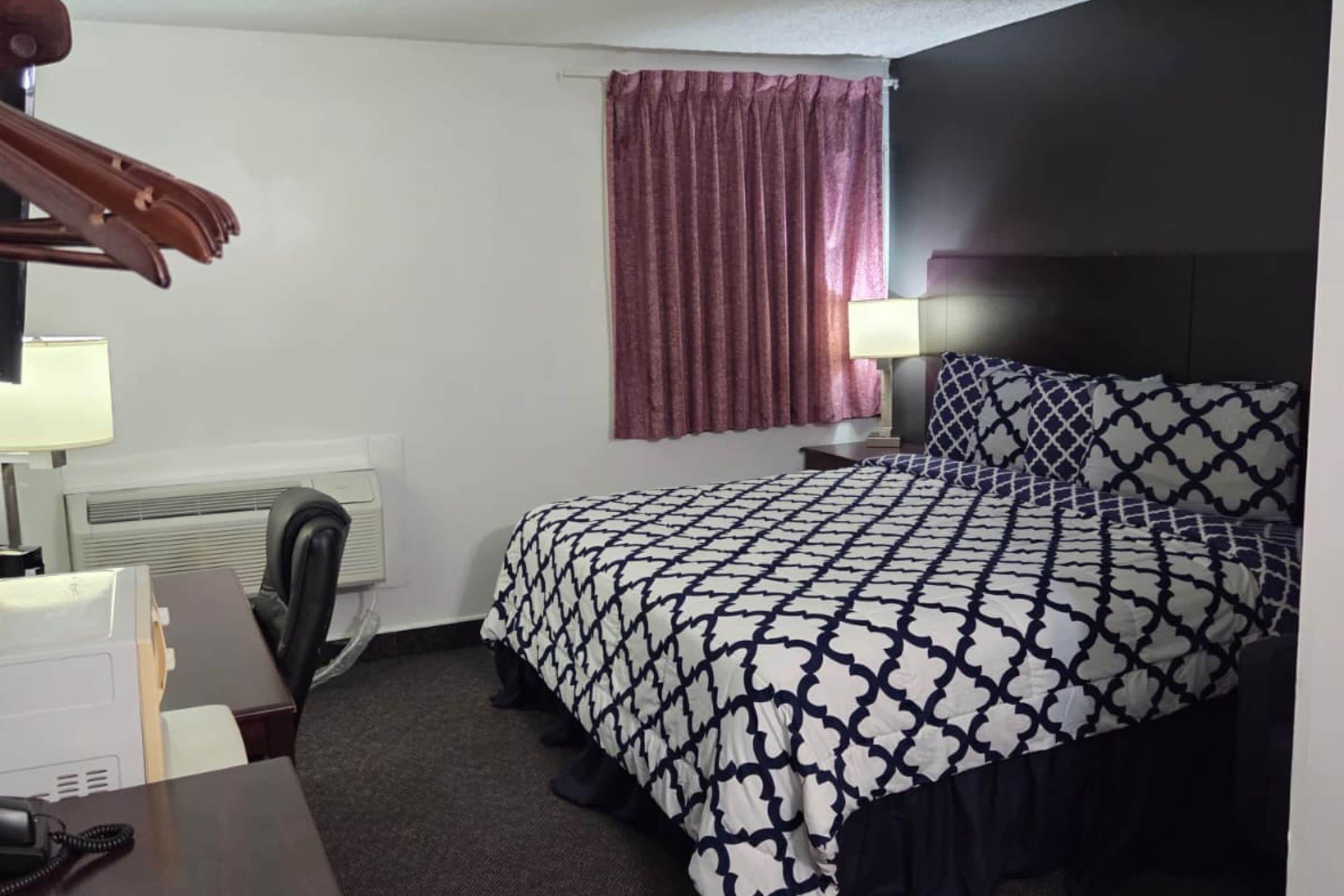 Habitación de hotel con cama, escritorio, silla, lámpara, calefacción y ventana con cortinas.
