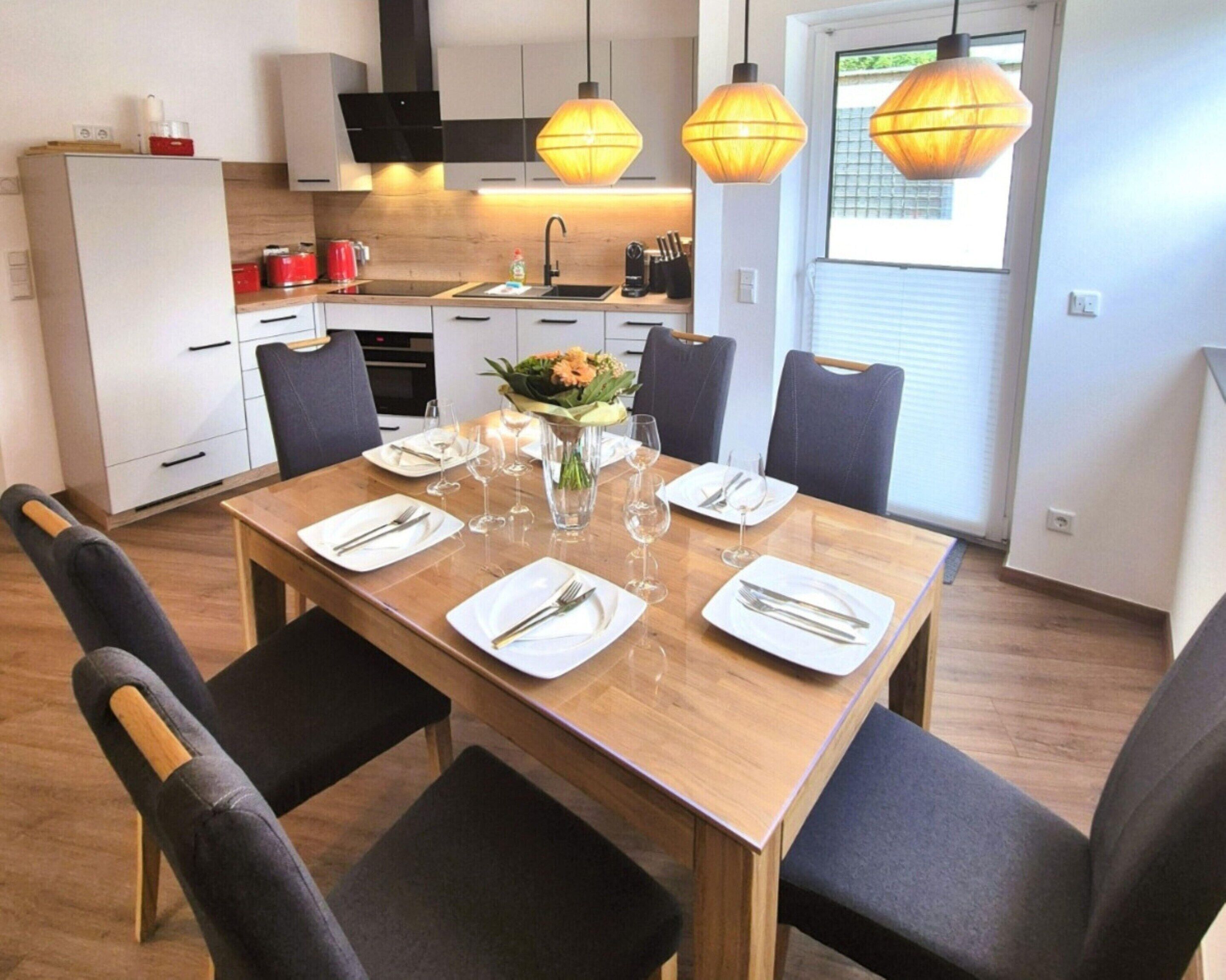 A modern kitchen with white cabinets, a wooden dining table set for four, and pendant lighting.