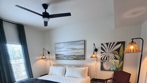 Ein Schlafzimmer mit einem Bett, einem Deckenventilator, einem Gemälde, zwei Wandlampen, einem Sessel und einem Teppich.