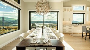 A modern dining room with a large table set for six, large windows offering a view of the mountains, and a kitchen with white cabinets and a large window. - Five-Star Base for Powder Runs & Lake Days - Adventure Elevated (Heber City)