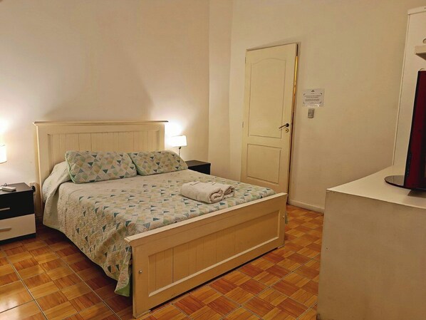 A bedroom with a wooden bed, a nightstand, a bedside lamp, and a door with a sign. - Hostel Estoril (Buenos Aires)