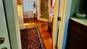 Un pasillo con piso de madera, un tapete estampado, un baño con puerta de vidrio para la ducha y un lavamanos.