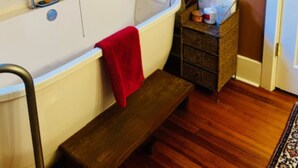 Un baño con bañera, un banco de madera, una pequeña mesita de mimbre con artículos de aseo y cuadros en la pared.