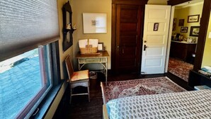 A bedroom with a bed, a chair, a desk, a window with blinds, a door, and a mirror. - The Burnt Toast Inn- Gold Room (Ann Arbor)