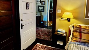 A hotel room with a wooden door, a bed with a plaid bedspread, a nightstand with a lamp, and a rug. - The Burnt Toast Inn- Gold Room (Ann Arbor)