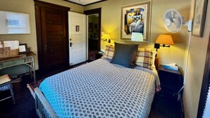 A hotel room with a bed, a desk, a chair, a standing fan, and a door leading to another room. - The Burnt Toast Inn- Gold Room (Ann Arbor)