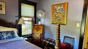 A bedroom with a bed, a bedside table, a lamp, a fan, a chair, a wooden chest, and a painting on the wall.