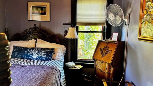 A bedroom with a bed, a window with blinds, a standing fan, a lamp, a dresser, and a framed picture on the wall.