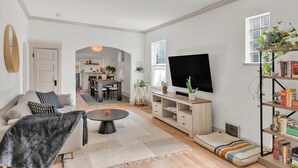 Ein großzügiger Wohnraum mit einer Couch, einem Couchtisch und einem Fernseher. Im Hintergrund ist eine Küche erkennbar, und ein Bücherregal beherbergt Bücher und Pflanzen.