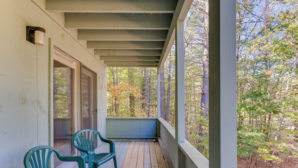 A covered porch with wooden flooring, two green plastic chairs, and a view of a forested area.