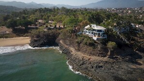 Una propiedad costera con una casa en un acantilado con vistas al mar, rodeada de vegetación y montañas a lo lejos.