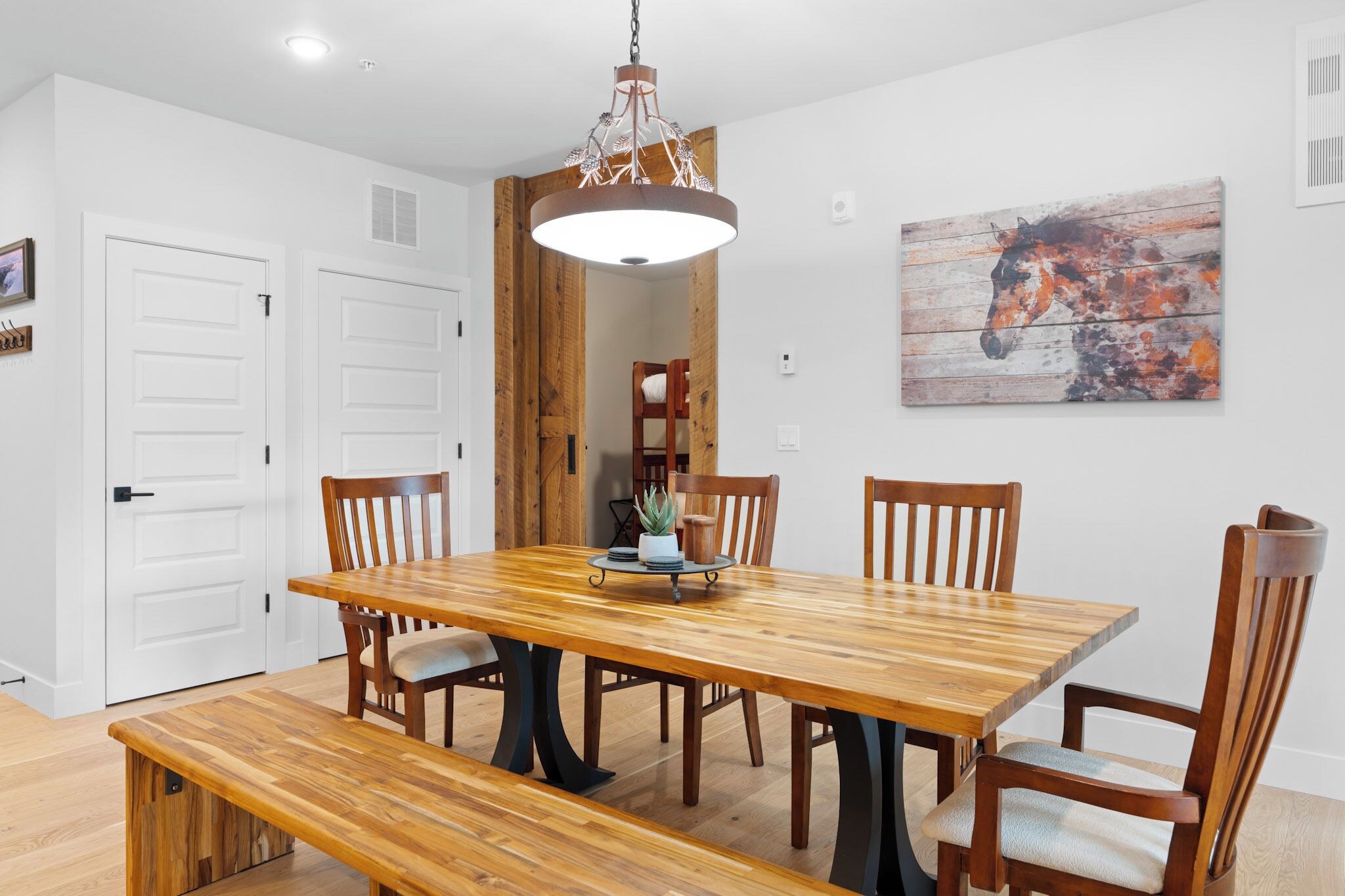 Un coin repas avec une table et des chaises en bois, un banc, un luminaire suspendu et un tableau sur le thème des chevaux accroché au mur.