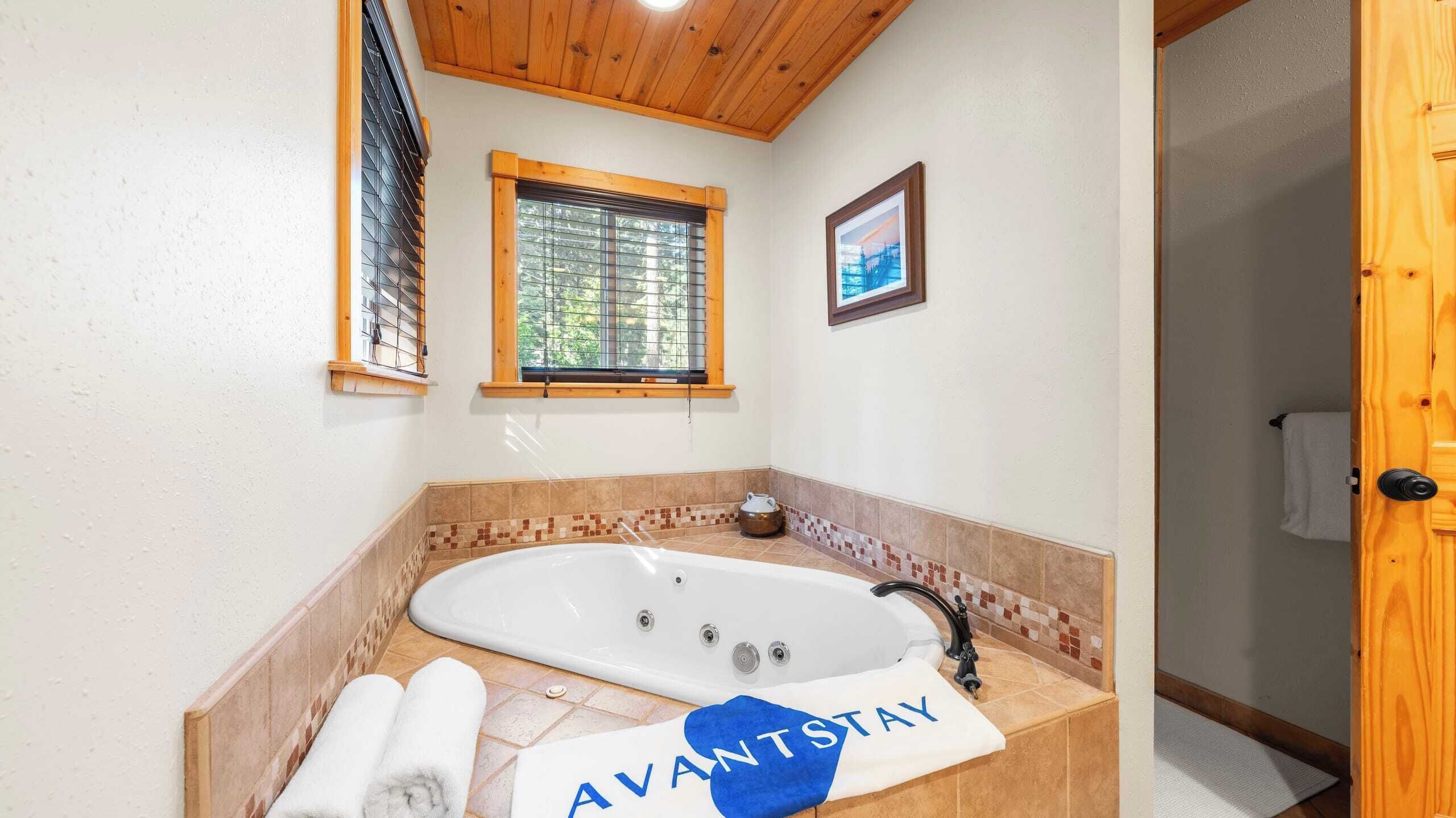 A bathroom with a large bathtub, a window with blinds, a framed picture on the wall, and a towel with "AVANTSTAY" on it.