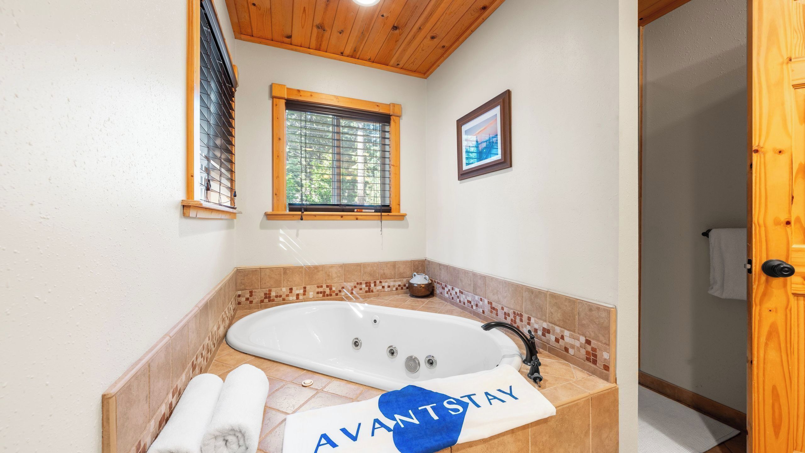 Un baño con una bañera grande, una ventana con persianas, un cuadro enmarcado en la pared y una toalla con 'AVANTSTAY'.