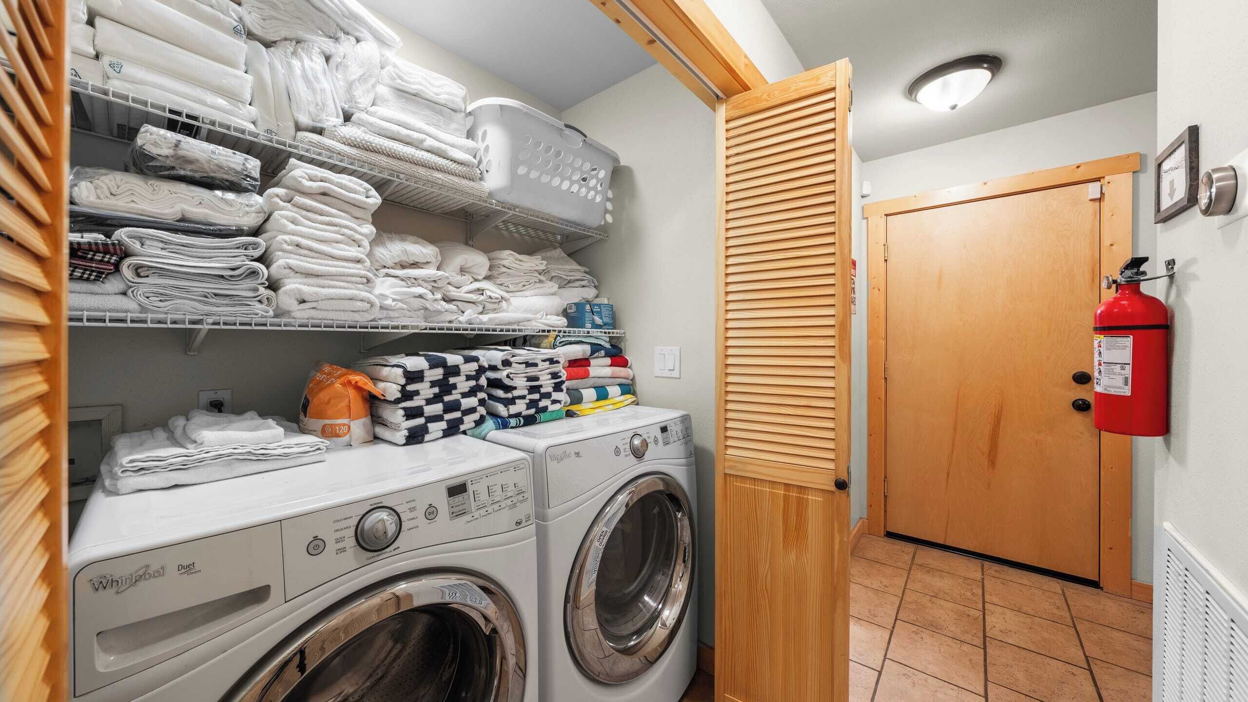 Ein Waschraum mit einer Whirlpool Duet Waschmaschine und einem Trockner, Regalen mit gefalteten Handtüchern und einem Feuerlöscher an der Wand.