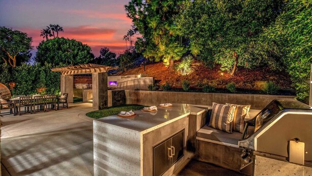 Une terrasse extérieure dotée d’un barbecue intégré et d’un espace salon, entourée d’une végétation luxuriante et agrémentée d’une structure en pierre avec un foyer.