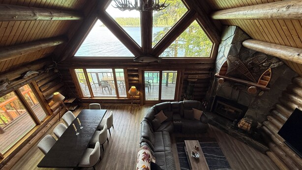 A log cabin interior with a dining area, sofa, fireplace, and large windows offering a view of the outdoors. - Ocean Front Property with Beach Access (Ketchikan)