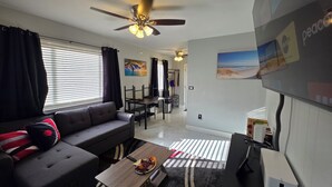 Una sala de estar moderna con un sofá, una mesa de centro y un ventilador de techo. Hay una zona de comedor con una mesa y sillas, y un televisor montado en la pared.