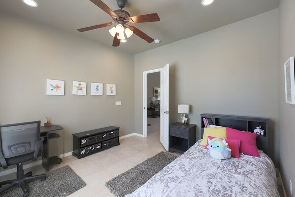 A bedroom with a bed, desk, and computer, a ceiling fan, and artwork on the wall.