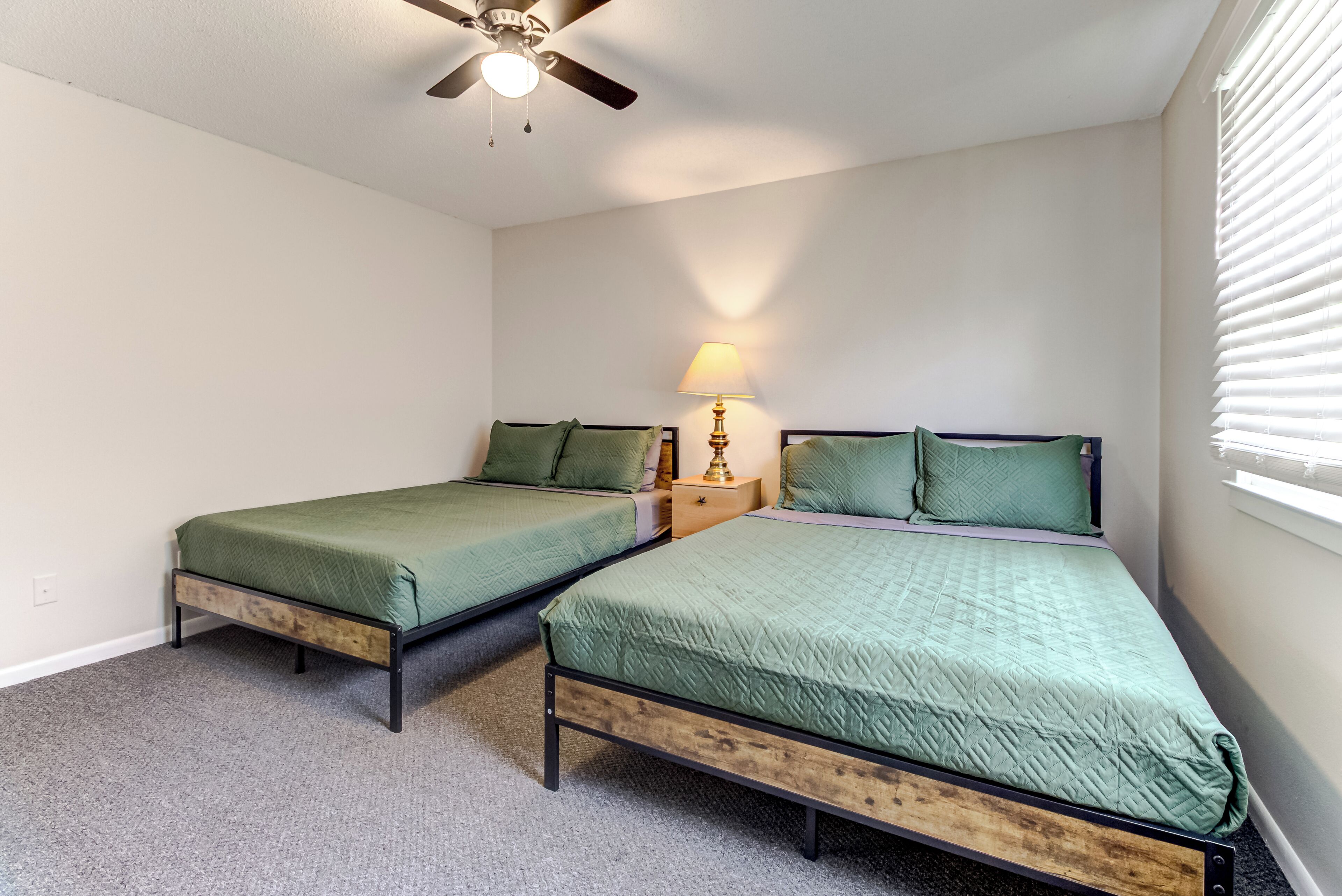A bedroom with two single beds, a ceiling fan, a bedside table with a lamp, and a window with blinds.