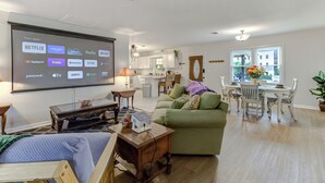 A modern living room with a large screen displaying streaming apps, a green sofa, a wooden coffee table, and a dining area with a white table and chairs. - Sunflower Theater - 3br, 2ba, 5 beds, 1600sqft with Smart Projector Movie Screen (Wilmington)