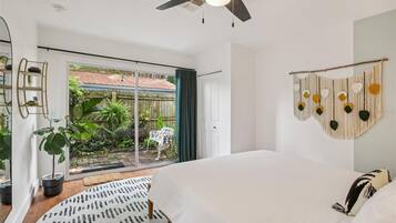 Un dormitorio con una cama grande, un ventilador de techo, vistas al jardín a través de una puerta corrediza y un tapiz de macramé en la pared.