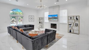 A spacious living room with a grey sectional sofa, a coffee table, and a fireplace. There is a large window with a view of greenery and a pool. - 7000sf Mansion w/ Pool/Gameroom/Theater - N (Davie)