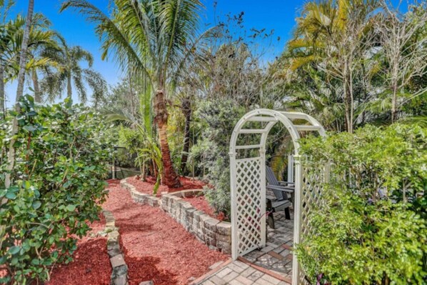 Un jardin agrémenté d’une pergola, d’un chemin recouvert de paillis rouge et de diverses plantes tropicales.