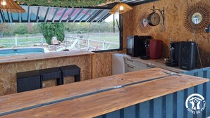 Een rustieke buitenduur bar met een houten toonbank, rieten stoelen en uitzicht op een omheinde tuin.