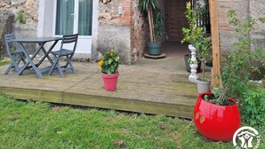 Een rustiek stenen huis met een houten terras, een overdekte veranda en potplanten.