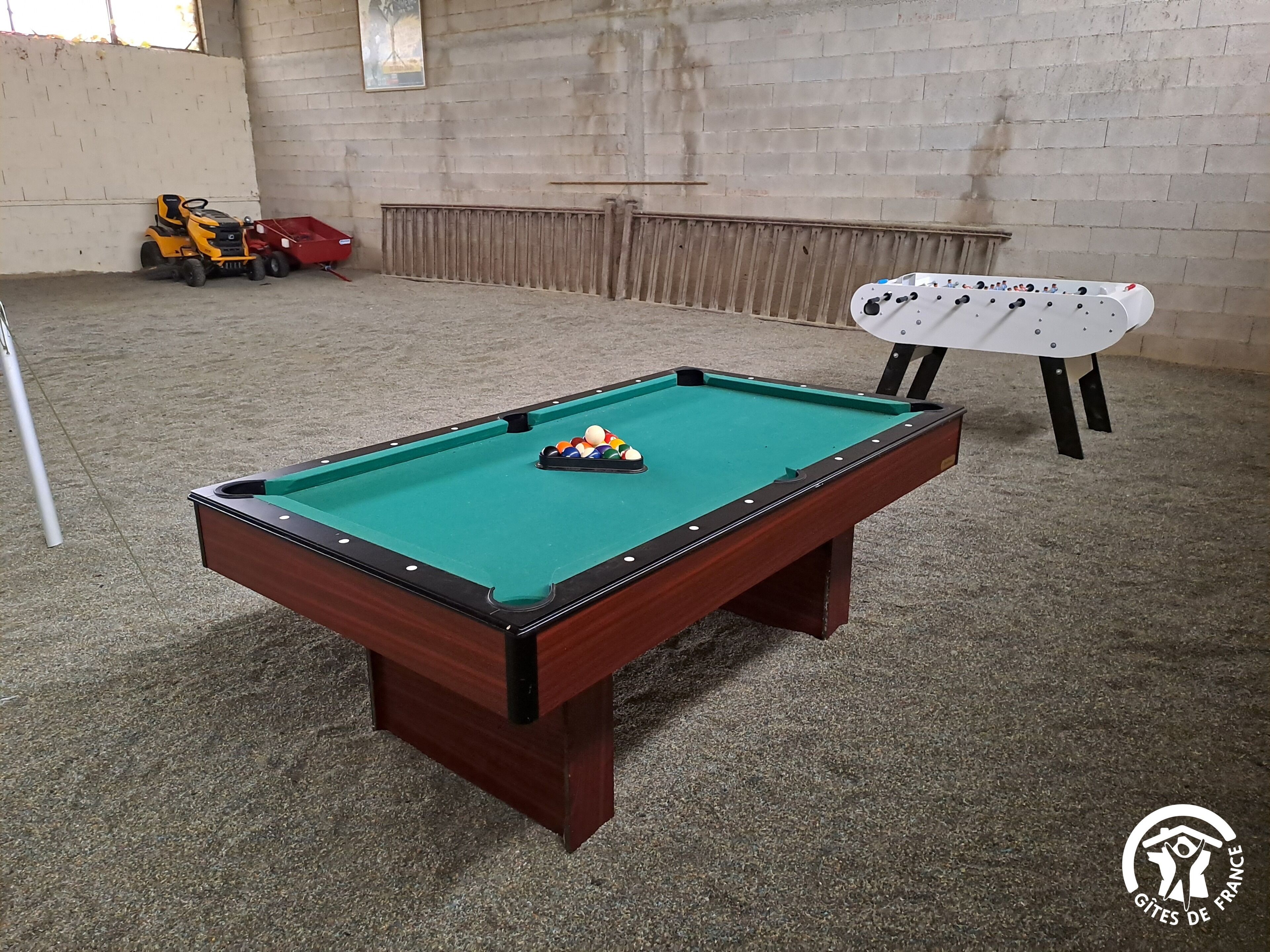 Een biljart met ballen in het midden, een rood speelgoedvoertuig, en een witte tafelvoetbaltafel in een ruimte met een betonnen vloer en bakstenen muren.