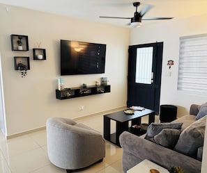 Un salon Ă©quipĂ© dâun tĂ©lĂ©viseur Ă Ă©cran plat, dâun ventilateur de plafond, dâune porte noire et dâun canapĂ© gris.