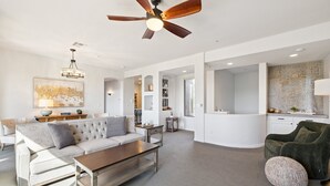 Un salón espacioso con un sofá grande, una mesa de centro y un ventilador de techo. Se ve una zona de cocina al fondo.
