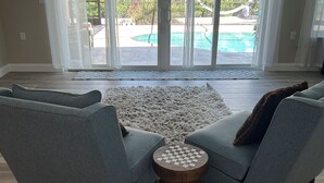 A living room with two grey armchairs, a round wooden side table, and a large glass door leading to a pool area.