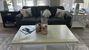 A living room with a grey sofa, two decorative pillows, a coffee table with a glass vase and seashells, a small side table with a lamp, and a pet bed in the corner.