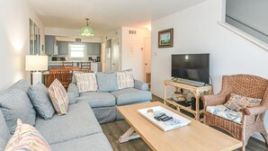 Una sala de estar moderna con un sofá seccional, una mesa de centro de madera, un sillón de mimbre, un televisor y una cocina al fondo.