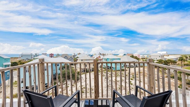 Una terraza de madera con dos sillas negras, con vista a una playa con casas coloridas y un cielo despejado.