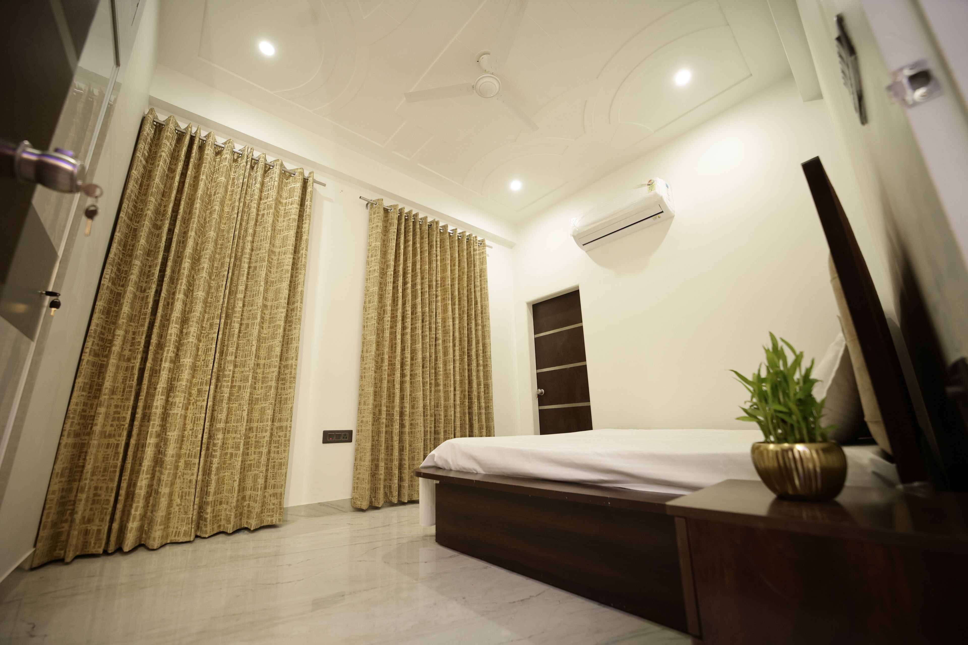Une chambre à coucher comprenant un lit, des rideaux, un appareil de climatisation, un ventilateur de plafond et une table de chevet agrémentée d’une plante.
