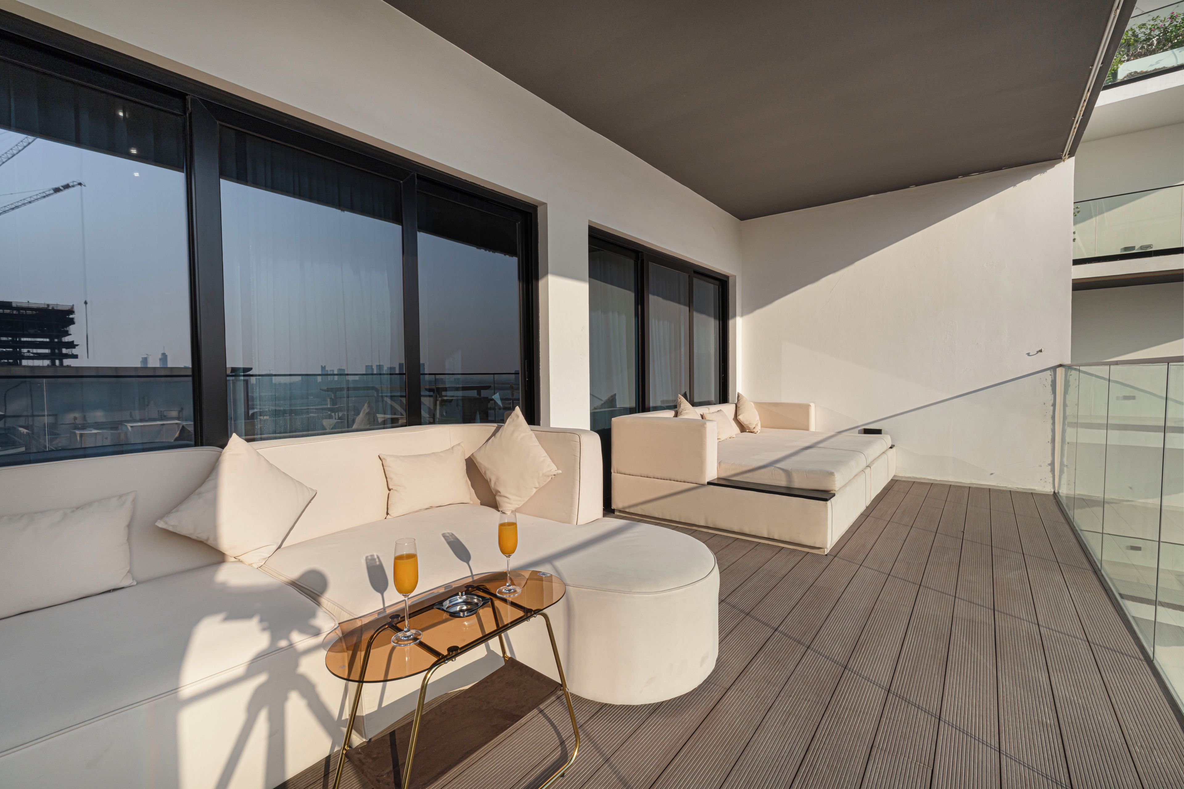 A modern outdoor seating area with a sofa and a glass table, a balcony with a view of the city skyline, and a wooden floor.