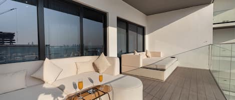 A modern outdoor seating area with a sofa and a glass table, a balcony with a view of the city skyline, and a wooden floor.