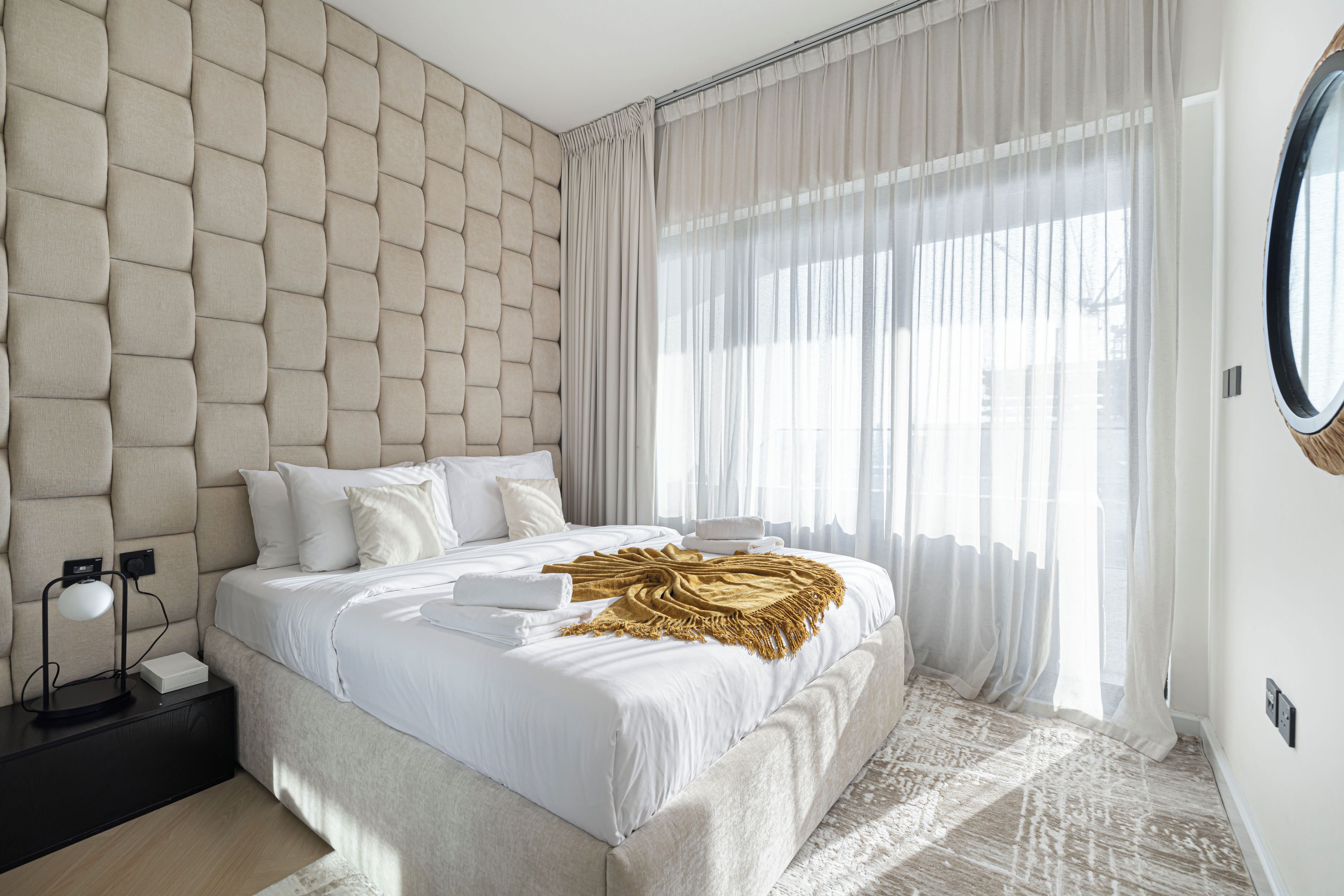 Une chambre à coucher moderne avec une tête de lit capitonnée, une grande fenêtre ornée de rideaux légers, une table de chevet avec une lampe, et un lit avec du linge de lit blanc et un plaid doré.