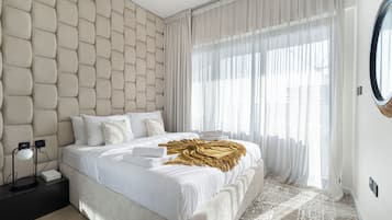 A modern bedroom with a tufted headboard, a large window with sheer curtains, a bedside table with a lamp, and a bed with white linens and a golden throw.