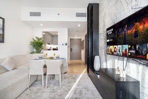 A modern living room with a dining area, a large TV screen displaying a series, and a view into the kitchen.