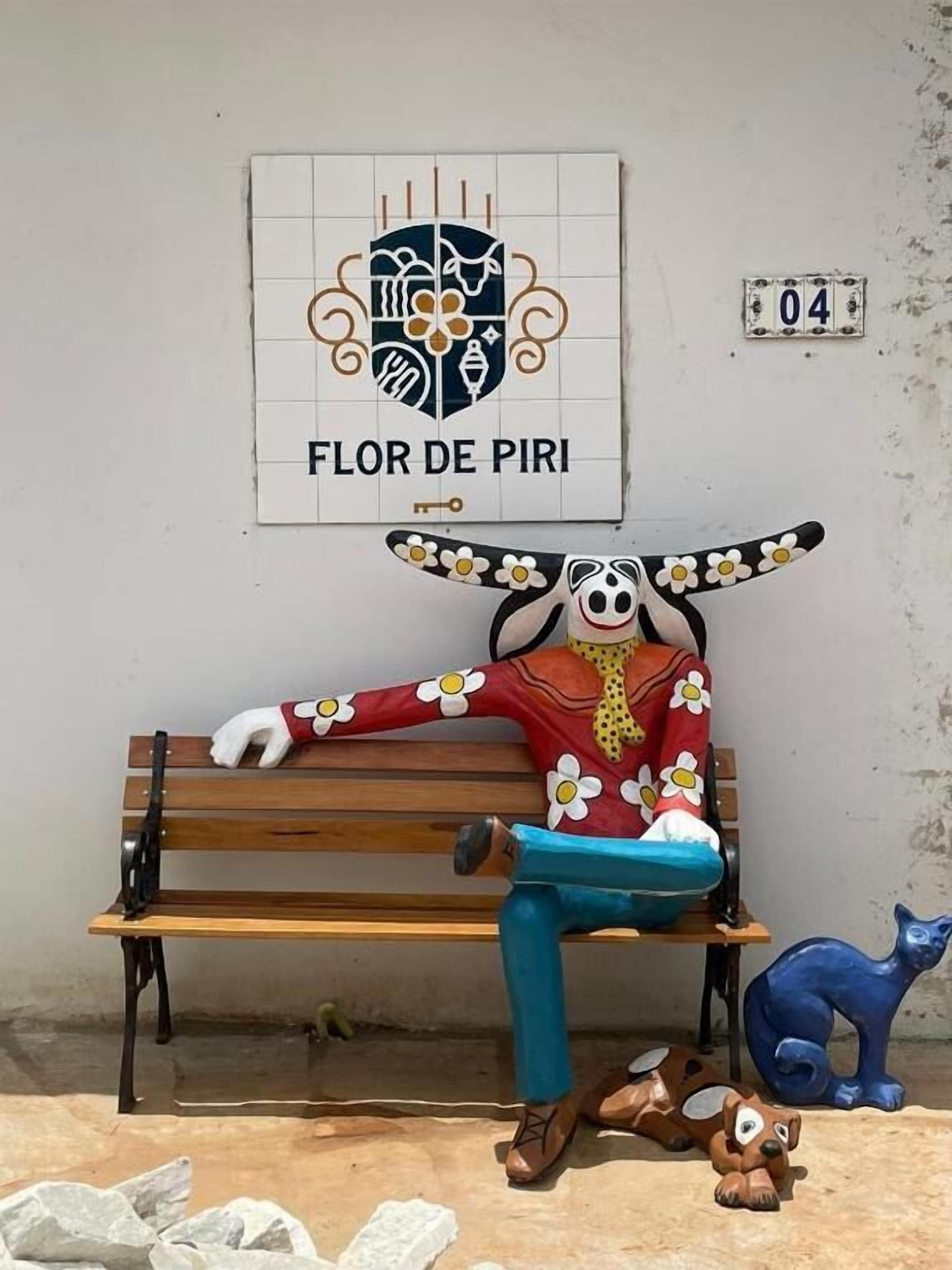 Un banc sur lequel est allongé une personne, un mur avec une pancarte indiquant « FLOR DE PIRI », et une statuette de chat bleue.