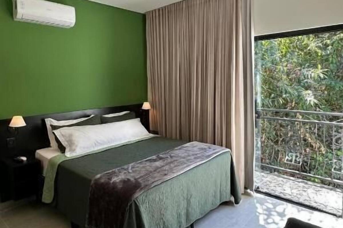 Un dormitorio moderno con cama, mesitas de noche, lámpara y vista a zonas verdes a través de una ventana.