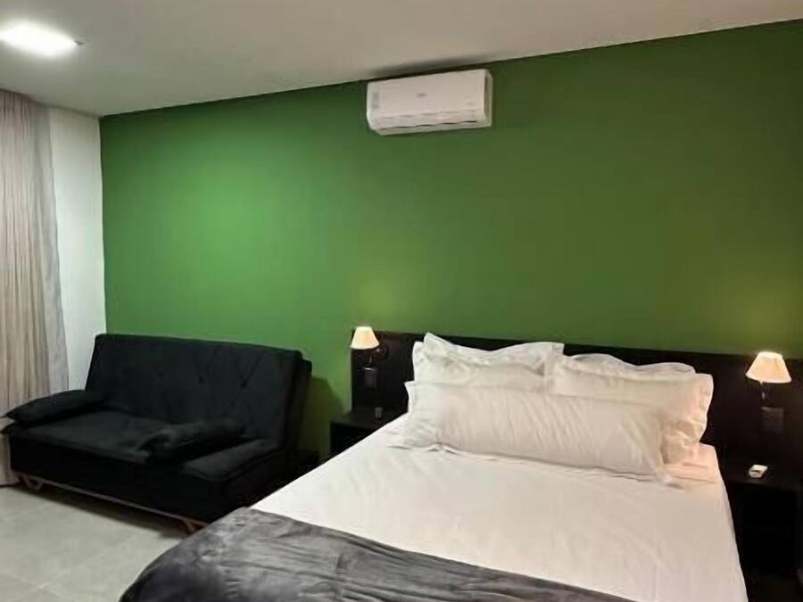 Une chambre d’hôtel avec un lit, deux tables de chevet avec des lampes, un mur d’accent vert, un canapé noir et un luminaire au plafond.