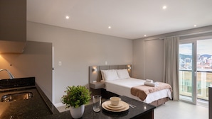 A modern hotel room with a bed, a small kitchen area, a dining table with a plant, and a view of the city through large windows. - Your Exclusive Stay in Blumenau, 350m From Vila Germanica - Ocns (Blumenau)