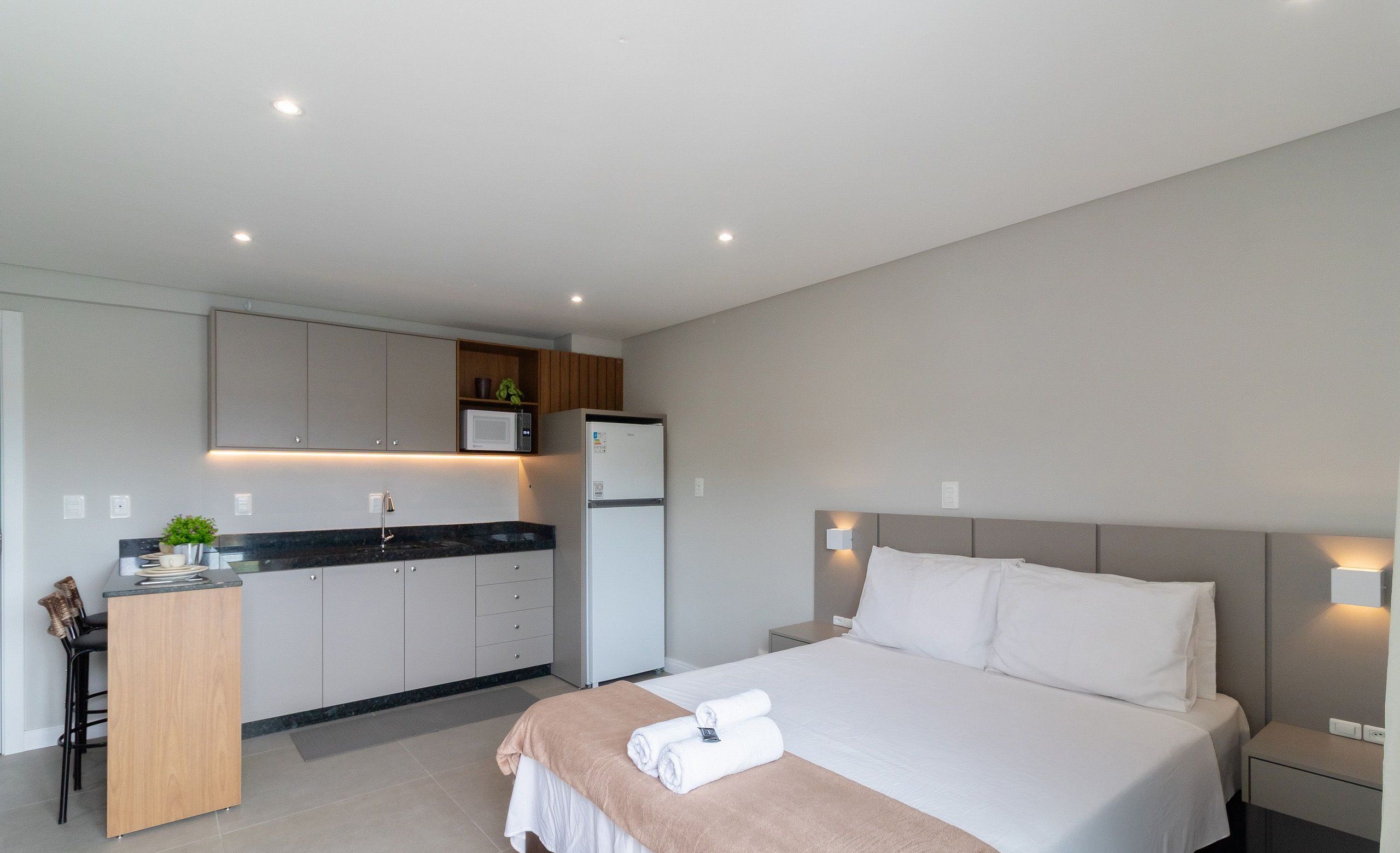 A modern hotel room with a bed, bedside tables, a desk, and a kitchenette.