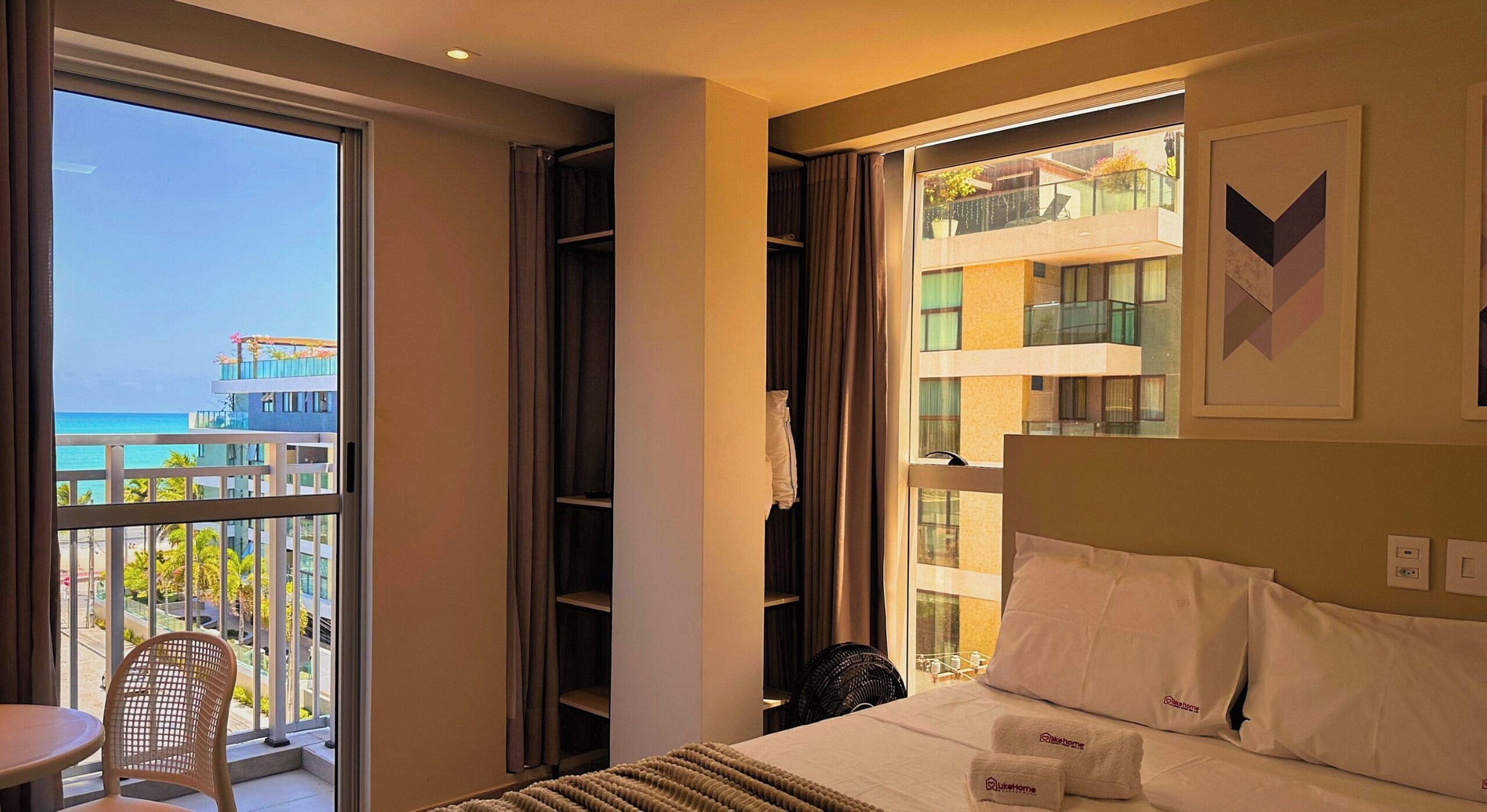 Quarto de hotel com cama, cadeira, ventilador e uma janela grande com vista para um edifício e uma varanda.
